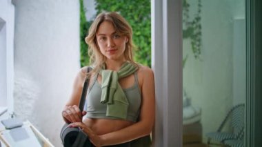 Kapalı mekanda sakin bir şekilde pencerenin yanında duran atletik kadın rahat bir şekilde yakından bakıyor. Yeşil spor kıyafetli genç bayan kameraya gülümseyerek poz veriyor. Kendine güvenen kızın portresi.