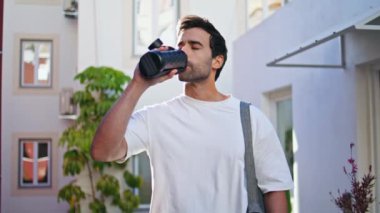 Modern mahallede siyah şişeden su içen sportif adam beyaz tişört giyiyor. Tıraşsız genç adam açık havada taze içecek yudumluyor. Aktif egzersiz rutininde susuzluk ve enerji.