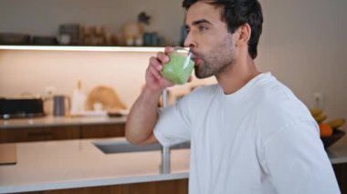 Modern mutfakta camdan yeşil smoothie içen bir adam. Gülümseyen İspanyol adam sağlıklı yaşam üzerine yoğunlaşarak taze vitamin kokteylinin tadını çıkarıyor. Dengeli yaşam tarzı için besleyici ev yapımı içecek.