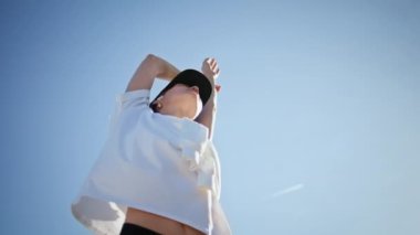 Açık havada eğitim gören esnek bir bayan mavi gökyüzünü yakından görebilmek için el kaldırıyor. Genç sporcu kadın kablosuz kulaklıklarla spor vücudunu esnetiyor. Sakin bir kadın doğa üzerinde egzersiz yapıyor..