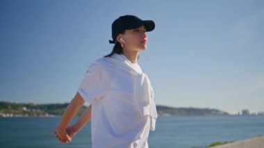 Sunny Street 'te esneyen, kablosuz kulaklıklar dinleyen bir kadın. Aktif kız nehir kıyısındaki spor eğitiminde vücut kaslarını ısıtıyor. Yaz rıhtımında şapkalı sporcu kadın egzersiz yapıyor.