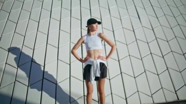 Kadın koşucu, şık spor kıyafetleri giyerek Sunny Street 'te antrenmanlara başladı. Atletik sporcu kadın sabah güneş ışığında geometrik duvarın yanında koşuyor. Modern şehir mimarisinin yakınında koşan aktif şapkalı bir bayan..