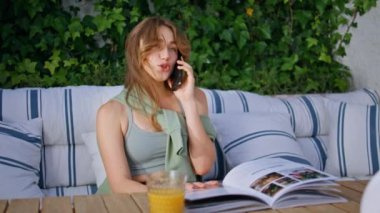 Sunlit apartman terasında akıllı telefondan konuşan şık bir öğrenci. Neşeli kadın rahat bir terasta canlı telefon konuşmasından hoşlanır. Mutlu kız cep telefonunu arıyor. Portakal suyuyla kafe masasında oturuyor..