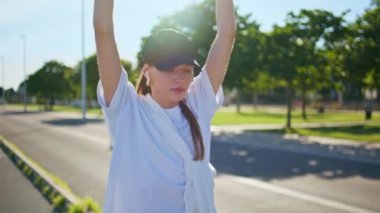 Yorgun koşucu, yaz sabahı koşu yaptıktan sonra dinlenirken yolda yürüyor. Sportif kadın elini kaldırıyor, koşudan sonra gözlerini kapatıyor. Sıska vücutlu sporcu kadın spor yapıyor..