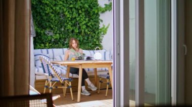 Etrafı yeşilliklerle çevrili, rahat bir terasta kitap okuyan genç bir kadın. Rahat bayan güneşli bir sabahta meyve suyu içerek sakin bir anın tadını çıkarın. Sayfaları çeviren güzel bir kız rahat bir koltukta oturuyor..