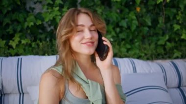 Gülümseyen kadın akıllı telefondan konuşuyor rahat kanepe terasında oturuyor. Rahat bir kafe terasında cep telefonu dedikodusu yapan mutlu bayan. Rahatlamış kız öğrenci sarmaşık yeşiliyle çevrili telefon görüşmesinin tadını çıkar..