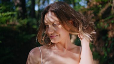 Parkta gün ışığını ısıtan gülümseyen kadın mutlu bir şekilde kameraya bakıyor. Çevresi yeşilliklerle çevrili, doğa maskeli güzel bir bayan. Mutlu kız dokunuyor saç stiline Neşeleniyor güneşli bir günde 