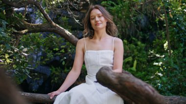 Orman kızı ağaçta tek başına oturup dinleniyor. Beyaz elbiseli, gülümseyen romantik bayan model ormanda şelalenin yanında rahatlayan kameraya bakıyor. Hayalperest kadın yeşilliğin tadını çıkarıyor doğa bahçeyi düşünüyor