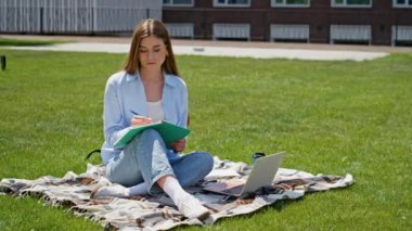Çimenli kampüs güneşli gününde düşünceli öğrenci notlar yazıyor. Genç bir kadın yeşil çim parkında oturan dizüstü bilgisayarla öğrenmeye odaklanmış. Güzel kız defteri kapatıyor. Canlı doğa projesini bitiriyor.