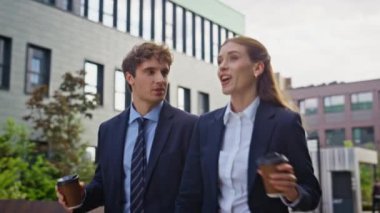 Neşeli iş ortakları ellerinde kahve fincanlarıyla sokakta yürüyorlar. Gülümseyen meslektaşlar modern kentsel alanda iş gezintisi yapıyorlar. İki iş arkadaşı şehir ortamında dostça sohbetin tadını çıkarıyor..