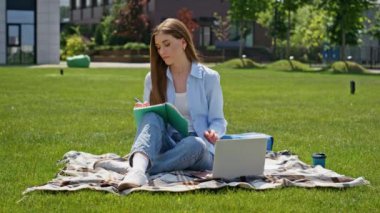 Çim kampüsünde okuyan genç bir kadın not defterine yazıyor. Sunny Park 'ta laptopla öğrenmekten zevk alan odaklanmış bir öğrenci. Yeşil çayır üzerinde tek başına oturan ciddi bir serbest çalışan kadın..