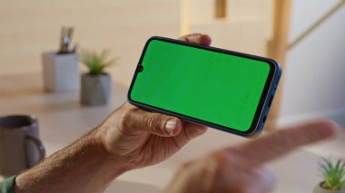 Modern iş yerinde krom tuşlu akıllı telefonu yakın planda tutan bir ofis çalışanı. Erkek elleri evde yeşil ekran cep telefonuna dokunuyor. Erkek serbest çalışan apartmandaki cep telefonundan fotoğraf izliyor..