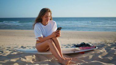Tatildeki bayan, Sandy Sahili 'nde akıllı telefonuyla güneş ışığını ısıtıyor. Huzurlu sörfçü kız arkadaşlarıyla mesajlaşıyor gezide dinleniyor. Güzel, rahat bir model sörf tahtasında dinleniyor. Seyahat hafta sonu konsepti