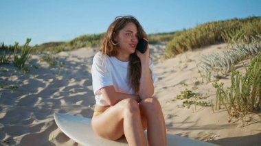 Plaj kızı haftasonu sörf tahtasında akıllı telefondan konuşuyor. Kumlu okyanus kıyısında dinlenen rahat bikinili kadın yaz gezisinde cep telefonunu arıyor. Sahilde iletişim kuran duygusal ve komik bir kadın.