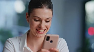 Cep telefonuyla sosyal medyayı izleyen neşeli bir kadın modern iş yerinde yakın çekim yapıyor. Ofis molasında gülümseyen iş kadını akıllı telefondan mesaj atıyor. Kapalı alanda esmer bir kadın cep telefonu mesajı okuyor..