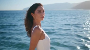 Şehvetli bayan deniz kenarında dinleniyor ve okyanus esintisinin tadını çıkarıyor. Güneşli okyanus sahilinde beyaz şık bir mayo giymiş gülümseyen bir kadın. Güneş ışığını yansıtan deniz dalgalarında poz veren güzel esmer.