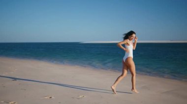 Deniz tatilinde sahilde güneşlenen bronzlaşmış bir bayan. Kaygısız, sıska bir kadın kumsala ayak basıp okyanus dalgaları saçıyor. Mutlu kız gezgin beyaz mayo giyerek güzel bir kıyı şeridinde eğleniyor..