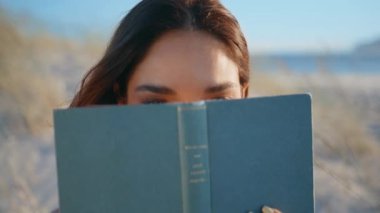 Okyanus sahilinde kitapların arkasına saklanan kadın gözleri. Portreli, çekici bir kadın. Kuru çimen deniz kıyısında oturuyor. Rahat gezgin güneşli sahil şeridinde eğleniyor.