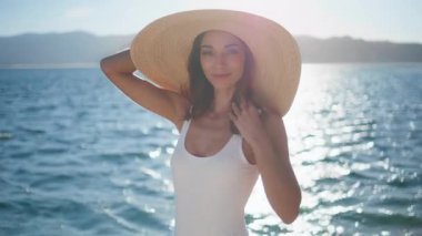 Lüks bir kadın okyanus kıyısında saman şapka takarak rahatlıyor. Parlak deniz suyu yüzeyinde yaz güneşi pozu veren şık bir model. Beyaz mayo giymiş, tropikal plajda duran mutlu turist kız..
