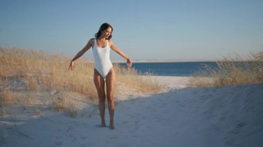 Okyanus kıyısının tadını çıkaran kaygısız kadın altın kum üzerinde dönüyor. Mutlu esmer, mavi gökyüzüne el kaldırıyor. Huzuru hissediyor. Güzel okyanus sahillerinde beyaz mayoyla yürüyen rahat bir kadın..