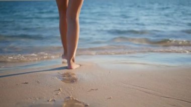 Yaz tatilinde ıslak kumsala adım atan bacaklar. Beyaz mayo giyen rahat bir kadın turist okyanus dalgaları üzerinde yürüyor ve kendini kaygısız hissediyor. Mutlu tatilciler güneşli deniz kıyısında geziniyor..