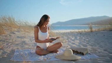 Plaj battaniyesinde açık bir kitap. Güzel kumlu sahil şeridinde rahatlamış esmer kadın romanı okuyordu. Gülümseyen bayan yaz tatilinde boş deniz kıyısında rahatlarken hobisinden zevk alıyor. Tasasız dinlenme konsepti