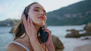 Kulaklıklı kız okyanus kıyısında dikiliyor ve yakın çekimin keyfini çıkarıyor. Gülümseyen güzel bir kadın yaz kumsalında müzik dinliyor. Mutlu kadın gezgin kayalık sahil doğasında dinleniyor..