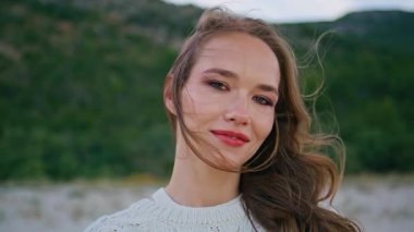 Yakından bakan kadın, doğada dinleniyor. Kameraya bakıyor ve gülümsüyor. Yeşil tepelerin önünde duran güzel genç bir kadının portresi rüzgarlı bir akşam. Rüzgarlı saçlı güzel manken sakin alacakaranlığın tadını çıkarıyor..