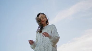Mavi gökyüzünün önünde poz veren kadın alt manzaraya yakın çekim yapıyor. Kaygısız kadın açık havada gökyüzünün altında gülümsüyor. Neşeli manken rahat beyaz kazak giyiyor ve rüzgarlı alacakaranlığın tadını çıkarıyor. Akşam doğasında dinlenen kız.