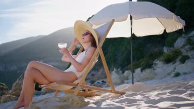 Plajda kokteyl içen bayan hasır şapkayla dinleniyor. Kumlu deniz kıyısında içki içen zarif genç bir kadın. Güneş gözlüklü mutlu esmer tatilde kaygısız hissediyor..
