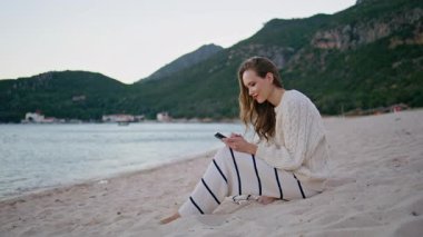 Sahil şeridinde cep telefonunu kaydıran kadın okyanus dalgalarının yanında sarı kumda oturuyor. Kumsaldaki akıllı telefondan sosyal medya izlerken gülen mutlu kadın. Gülümseyen kız turist deniz tatilinde telefon mesajı okuyor..
