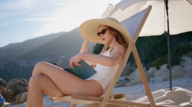 Kumsalda kitap okuyarak güneşlenmenin tadını çıkaran mutlu bir tatilci. Tasasız kız okyanus kıyısında rahatlarken şık güneş gözlüklerini çıkarıyor. Hasır şapka giyen güzel bir kadın sahilde okumayı bitiriyor..