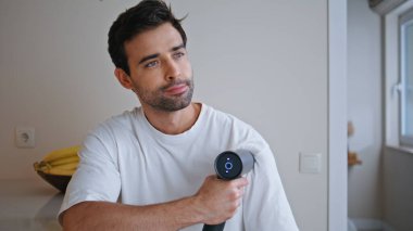 Omzunda elektronik masaj tabancası olan beyaz t-shirt giyen bir adam. İspanyol atlet antrenmandan sonra perküsyon cihazıyla kaslara masaj yapıyor. Eğitimden sonra iyileşen adam..