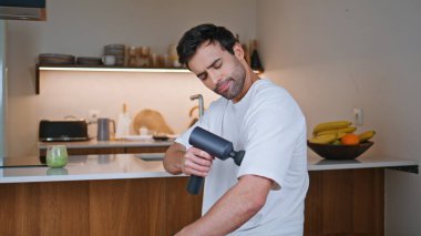 Sporcu omuz omuza masaj tabancası uyguluyor modern mutfağa yakın çekim yapıyor. Latin bir adam elektronik perküsyon masaj aletini kullanarak kas ağrısını hafifletir. Spor sonrası kişisel bakım eğitimi..