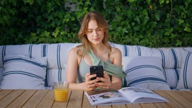 Gülümseyen kız cep telefonunun içeriğine bakıyor açık kafede oturuyor. Yeşil sarmaşık yapraklarıyla çevrili koltukta cep telefonu mesajı okuyan mutlu kadın. Sosyal medya akıllı telefonu izleyen hoş bir bayan.