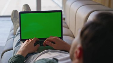 Freelancer oturma odasındaki kanepede çevrimiçi maket dizüstü bilgisayarda çalışıyor. Kimliği belirsiz bir adam sosyal medyada yeşil ekran bilgisayarına bakıyor ve kanepesinde yatıyor. Genç adam dairede krom anahtar kullanıyor.
