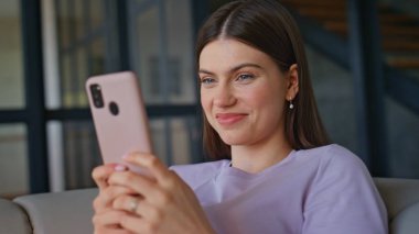 Modern apartman dairesinde cep telefonuna mesaj yazan ev kızı. Akıllı telefondan sosyal medyada gezinen gülümseyen genç bir kadın. İnternetten alışveriş yapan neşeli bir bayan oturma odasındaki koltukta cep telefonu kullanıyor. 