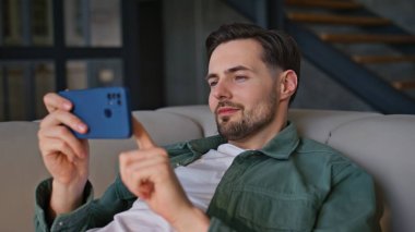 Akıllı telefon kullanan sıradan bir hippi kanepeye yakın çekim yapıyor. Hafta sonları rahat bir oturma odasında gülümseyen esmer cep telefonu. Cep telefonu tutan olumlu bir adam içerideki komik video içeriğinin keyfini çıkarıyor