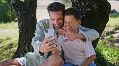 Gülümseyen baba oğul modern akıllı telefonu kullanarak güzel doğayı görüntülüyor. Mutlu baba, el sallayarak dışarıdaki cep telefonu kamerasına bakıyor. Parkta neşeli aile blogları 