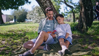 Baba çocuk ormanda yeşil çimlerde otururken kitap izliyor. Sevimli küçük çocuk, hafta sonunu babasıyla büyük bir ağacın altında geçirerek mutluluğunu ifade ediyor. Peri masalı okuyan anne babanın sevimli ailesi. 