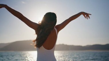 Güneş ışığı plajında gerinen beyaz mayo giymiş bir kadın. Sıska esmer kız, parlak akşam güneşinin tadını çıkarıyor. Kaygısız gezgin güzel okyanus dalgalarında tek başına duruyor..