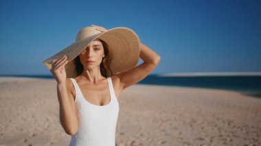 Yakın plan hasır şapka giyen şık bir model plaja poz veriyor. Beyaz mayo giymiş muhteşem bir kadın kumlu sahil şeridi kamerasına bakıyor. Zarif esmer deniz tatilinde yaz güneşinin tadını çıkarıyor..