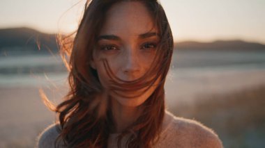 Mutlu esmer, gün ışığında ayakta duruyor ve gökyüzüne el kaldırıyor. Doğaya poz veren gülümseyen kadın altın günbatımı kamerasına bakıyor. Tasasız portre rüzgarlı manzarada mutluluğu hisseder..