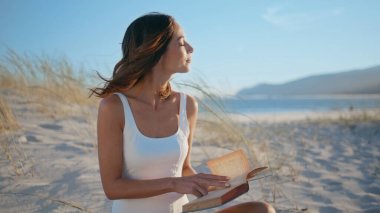 Sakin kız rüzgârlı kıyı şeridi hakkında kitap okuyor. Yaz haftasonu kumlu okyanus kıyısında oturup sayfaları çeviren gülümseyen kadın. Kaygısız esmer edebiyat hobisinin keyfini çıkarıyor güneşli deniz kenarında dinleniyor..