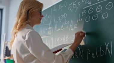 Öğretmen, tahtaya matematik denklemleri yazıyor. Kitaba yakından bakıyor. Karatahta sınıfındaki matematik ödevlerini çözen kadın eğitimci. Lisede cebir dersi veren, gözlüklü sarışın bir öğretmen..