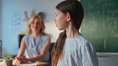 Küçük liseli kız karatahtaya yakın çekimde cevap veriyor. Genç bayan öğretmen sınıf tahtasında konuşan ilkokul çocuğuyla etkileşim içinde. Bayan eğitimci sınıfta ilkokul öğrencileriyle konuşuyor..