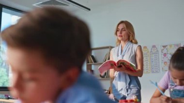 Öğrencileri gözlemleyen bir kitapla okul öğretmeni yürüyor. Kitaplara yazı yazan zeki okul çocukları sınıf sıralarını kapatıyorlar. İlkokulda çocuklara yardım eden kadın eğitimci.