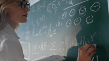 Öğretmen el yazısı formülleri karatahta okulunda yakın çekim. Gülümseyen kadın sınıfta matematik teorisi öğretiyor. Bilinmeyen bir öğrenciyle konuşan, sınıf dersinde matematik denklemlerini açıklayan bir kadın eğitmen.