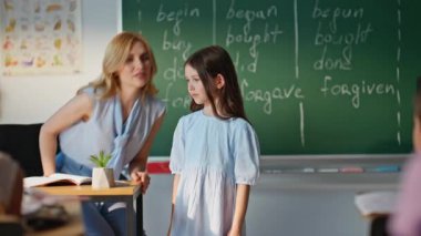 Karatahtadaki ilkokul öğrencisi öğretmen sorusuna cevap veriyor. Küçük liseli kız, yazı tahtasının yanındaki konuşmadan sonra masaya oturur. Genç bayan eğitmen ilkokul çocuklarıyla konuşuyor.