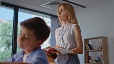 Odaklanmış öğrenciler sınıfa yakın çekimde notlar yazıyorlar. Kadın öğretmen elinde ilkokul öğrencilerini sınıftaki eğitim aktivitesinde gözlemleyen bir kitap tutuyor. Akademik adanmışlıkla çalışan düşünceli çocuklar..
